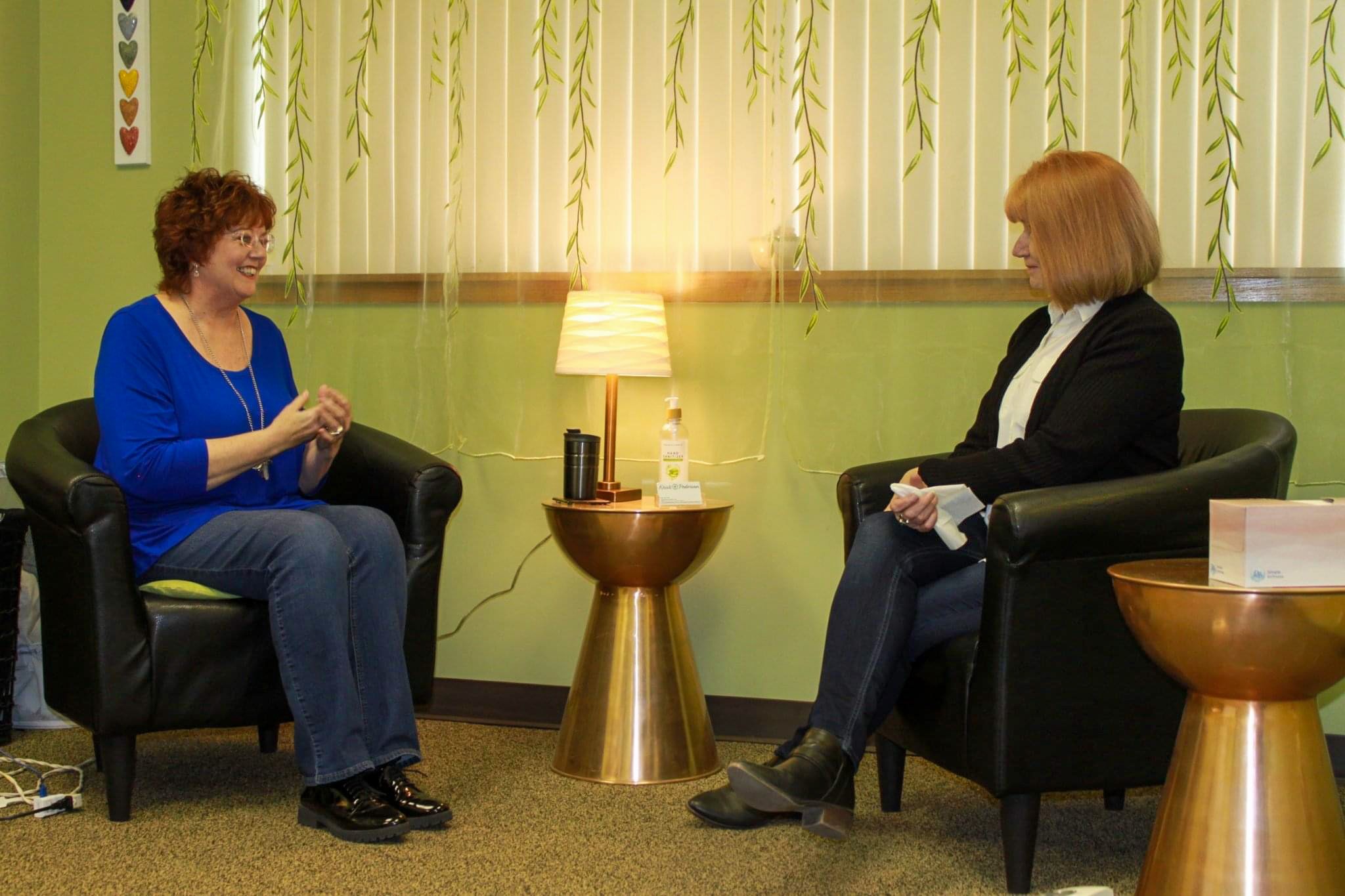 Kristi Pederson seated in a chair during a one-on-one conversation, speaking with another woman in a calm, welcoming studio setting.