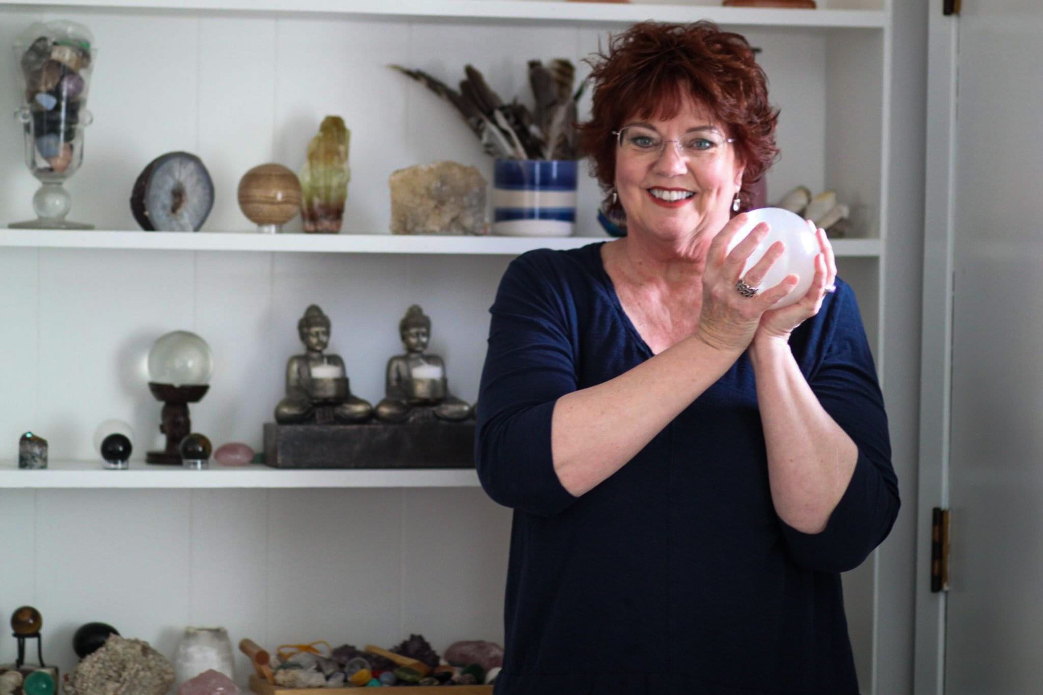 Kristi Pederson smiling while holding a glowing crystal sphere, standing in front of shelves filled with spiritual objects and crystals.