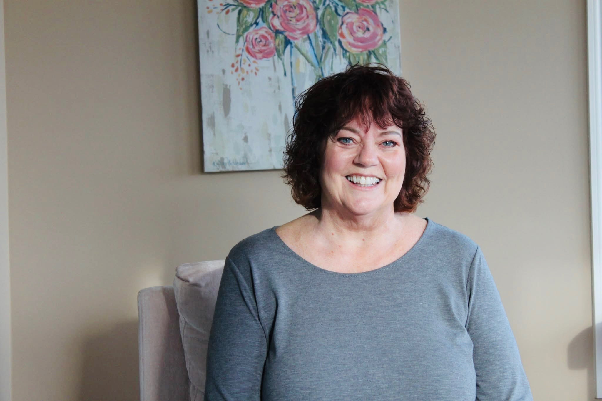 Kristi Pederson seated indoors, smiling warmly in a relaxed setting with artwork displayed behind her.