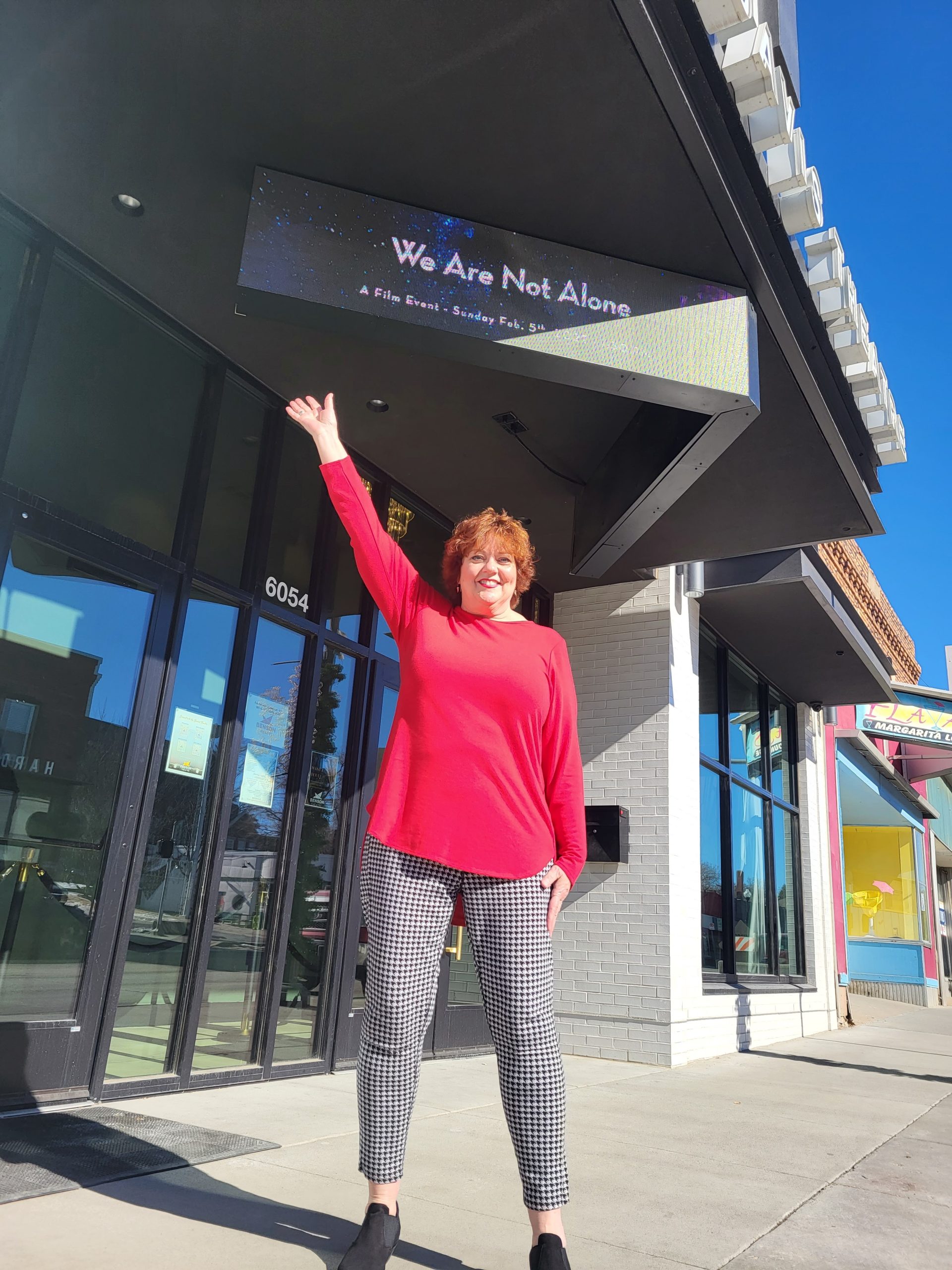 Kristi Pederson standing outside a storefront beneath a marquee sign, smiling and raising one arm in a welcoming gesture on a sunny day.